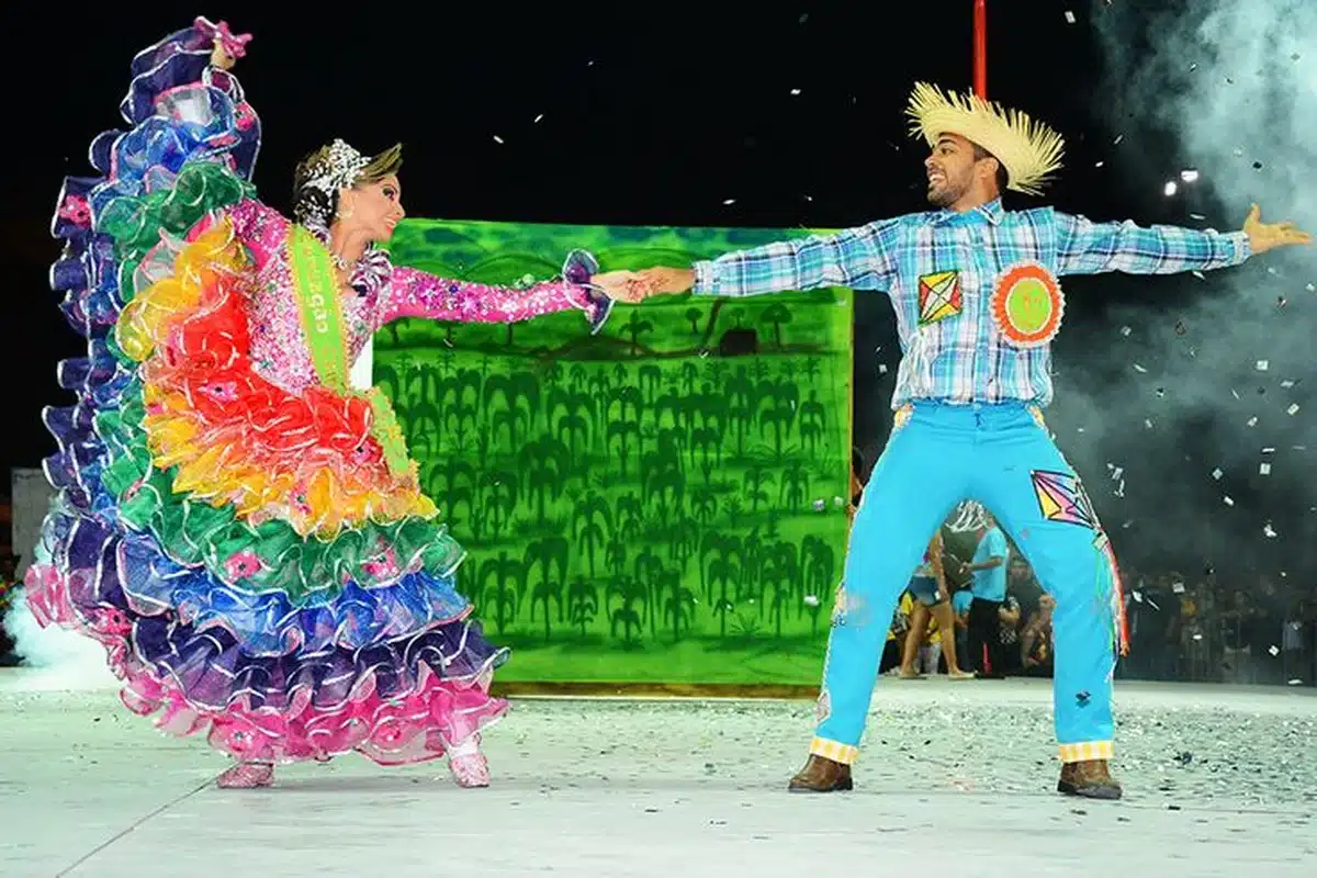Casal dançando quadrilha com trajes típicos de São João, com chapéu de palha e roupas coloridas.