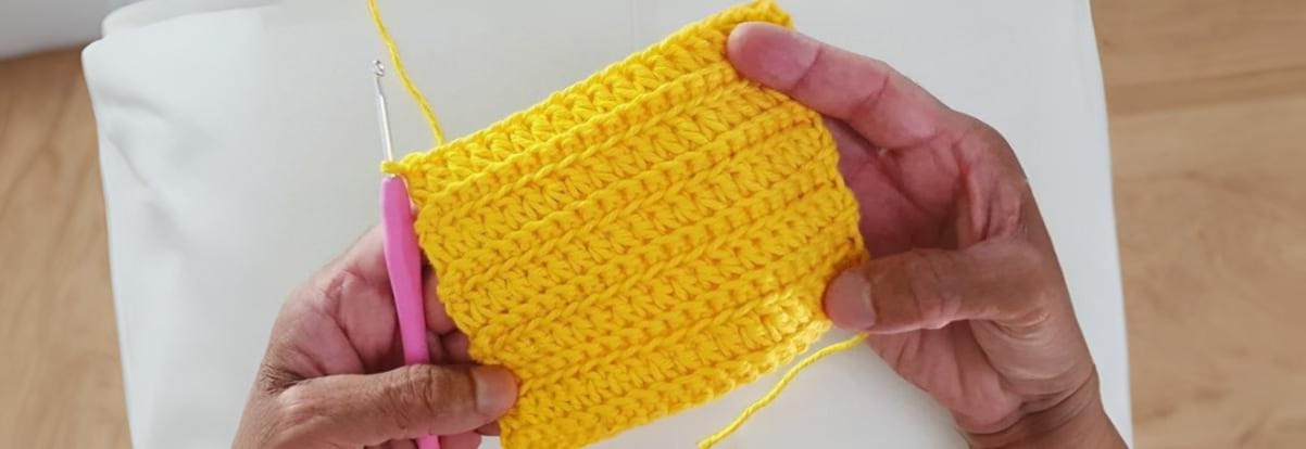 ãos segurando amostra de crochê amarelo vibrante com ponto texturizado, mostrando técnica de crochê fácil com agulha rosa e fio de algodão.