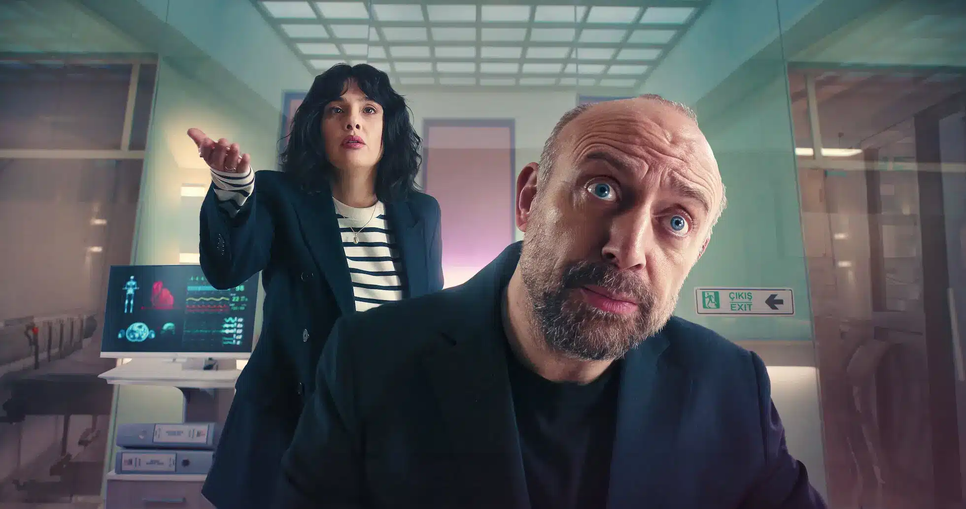 Cena do filme Amantes Anônimos (2025), com dois personagens em um cenário futurista, onde uma mulher expressa preocupação enquanto o homem parece surpreso, com gráficos médicos ao fundo.