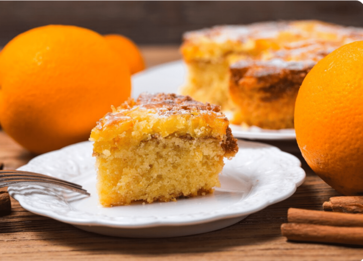 Fatia de bolo de laranja caseiro servida em prato branco com laranjas ao fundo