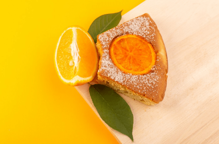 Fatia de bolo de laranja com fatia da fruta por cima, em fundo amarelo vibrante