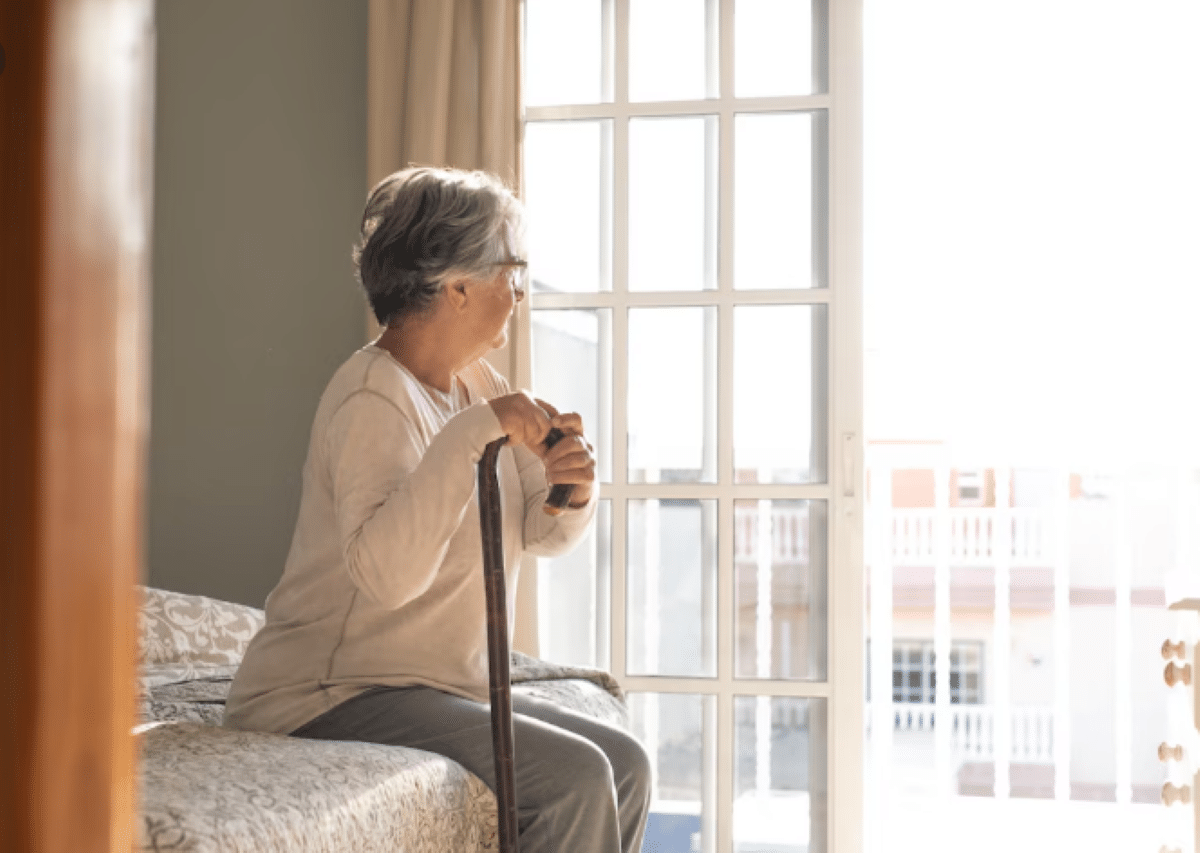 Idosa com bengala sentada na cama olhando pela janela em ambiente residencial