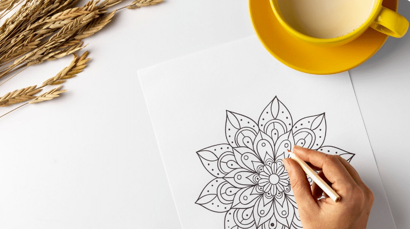 Mão colorindo mandala em folha branca com xícara amarela ao lado