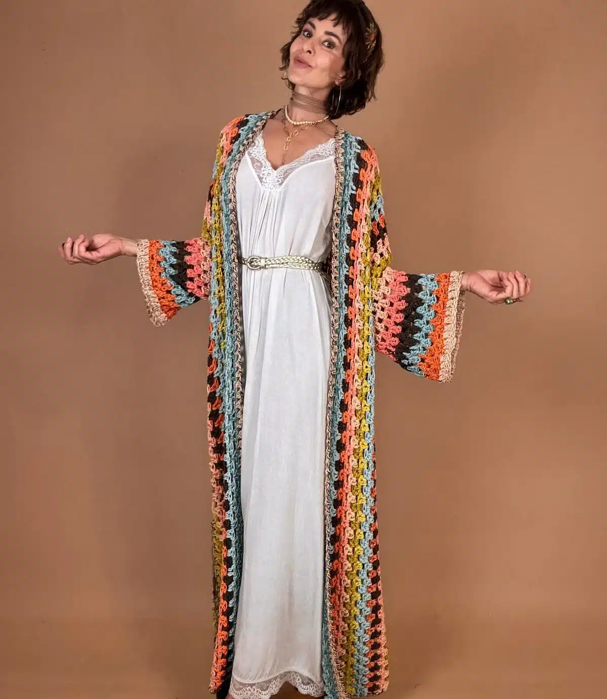Mulher usando um casaco kimono de crochê colorido com uma roupa branca e acessórios delicados, com um fundo neutro.