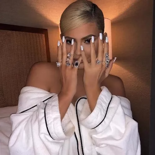 Kylie Jenner com esmalte branco e várias joias, posando com as mãos cobrindo o rosto