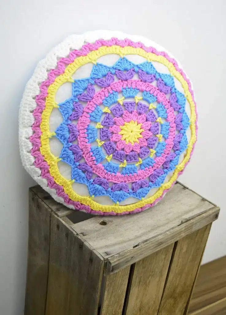 Almofada redonda com padrão de mandala colorida em tons de rosa, lilás, azul e amarelo.