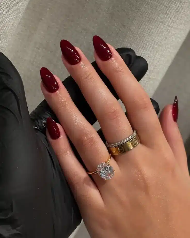 Unhas de gel vermelhas estilo stiletto com acabamento brilhante, combinadas com anéis de luxo dourado e prateado.