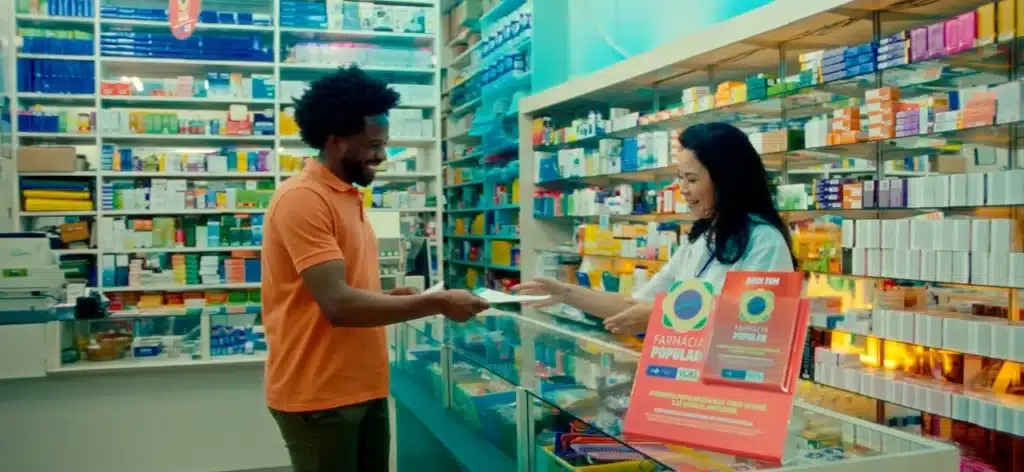 Homem sorridente entrega receita a farmacêutica no balcão, ilustrando o acesso facilitado a medicamentos pelo programa Farmácia Popular.