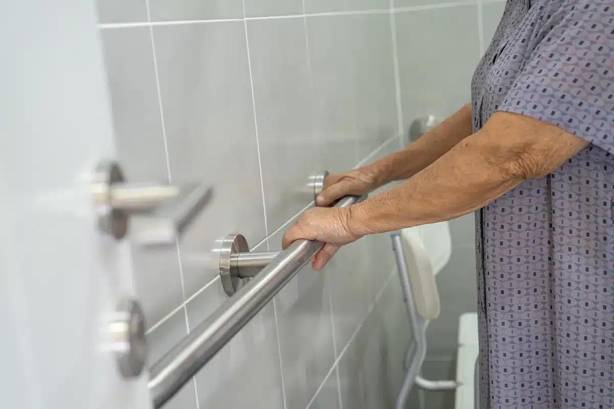 Pessoa idosa segurando barra de apoio instalada em banheiro adaptado.