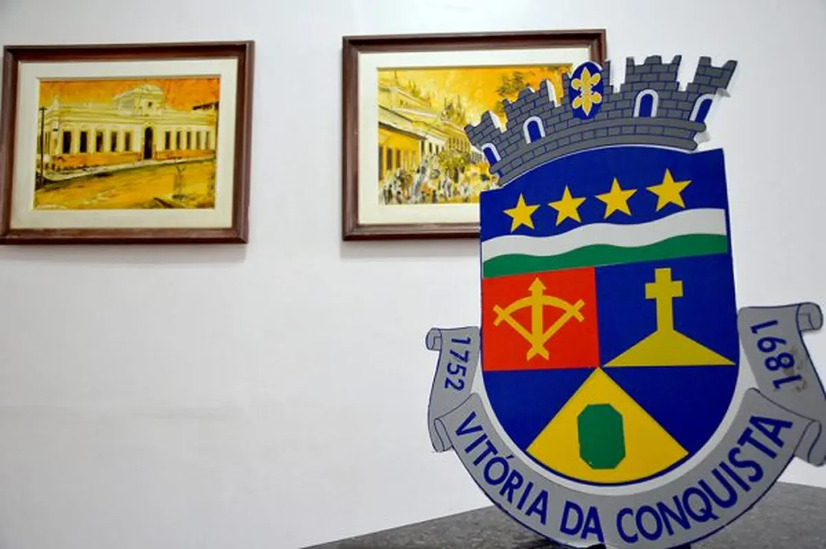 Brasão de Vitória da Conquista, cidade da Bahia, com detalhes de uma cruz, uma espada, e estrelas, no contexto de alerta sobre cadastro habitacional gratuito.