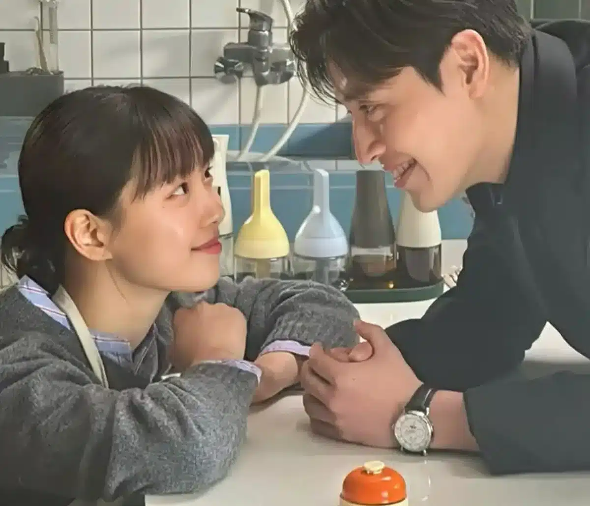 Cena de dorama com um casal em momento íntimo e carinhoso na cozinha, com expressão de afeto e cumplicidade.