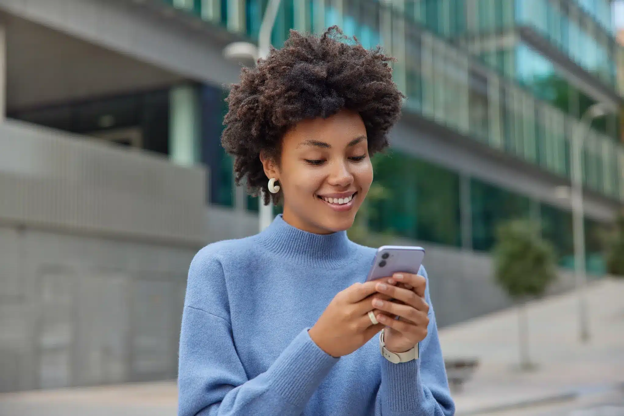 Jovem sorridente com cabelo cacheado, segurando o celular, transmitindo alegria e boas vibrações para um fim de semana incrível.
