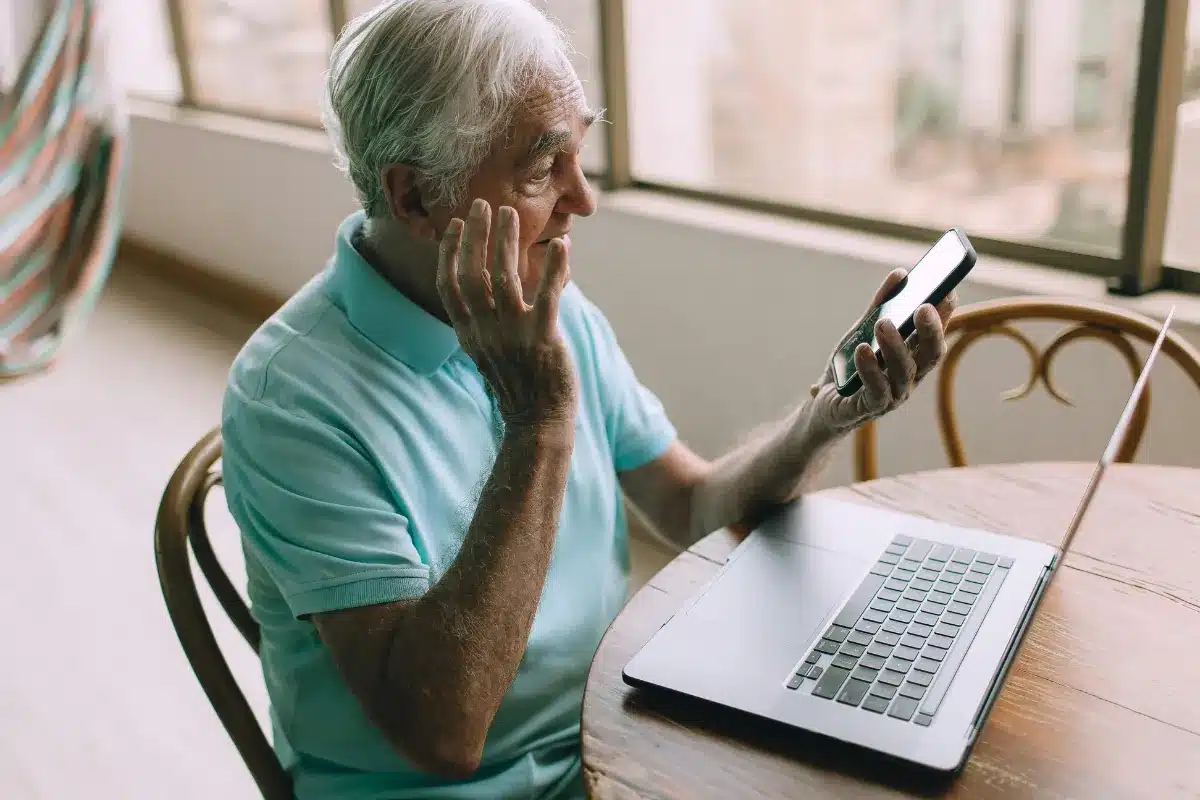 Idoso confuso com celular enquanto usa notebook.