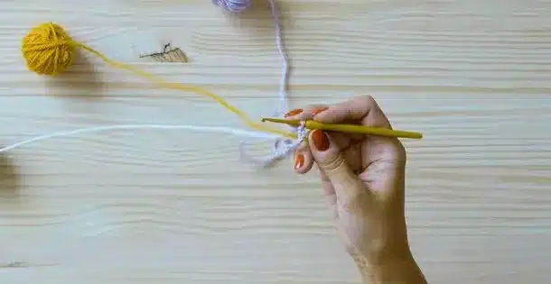 Mão segurando agulha de crochê ao iniciar ponto pipoca com três cores de linha