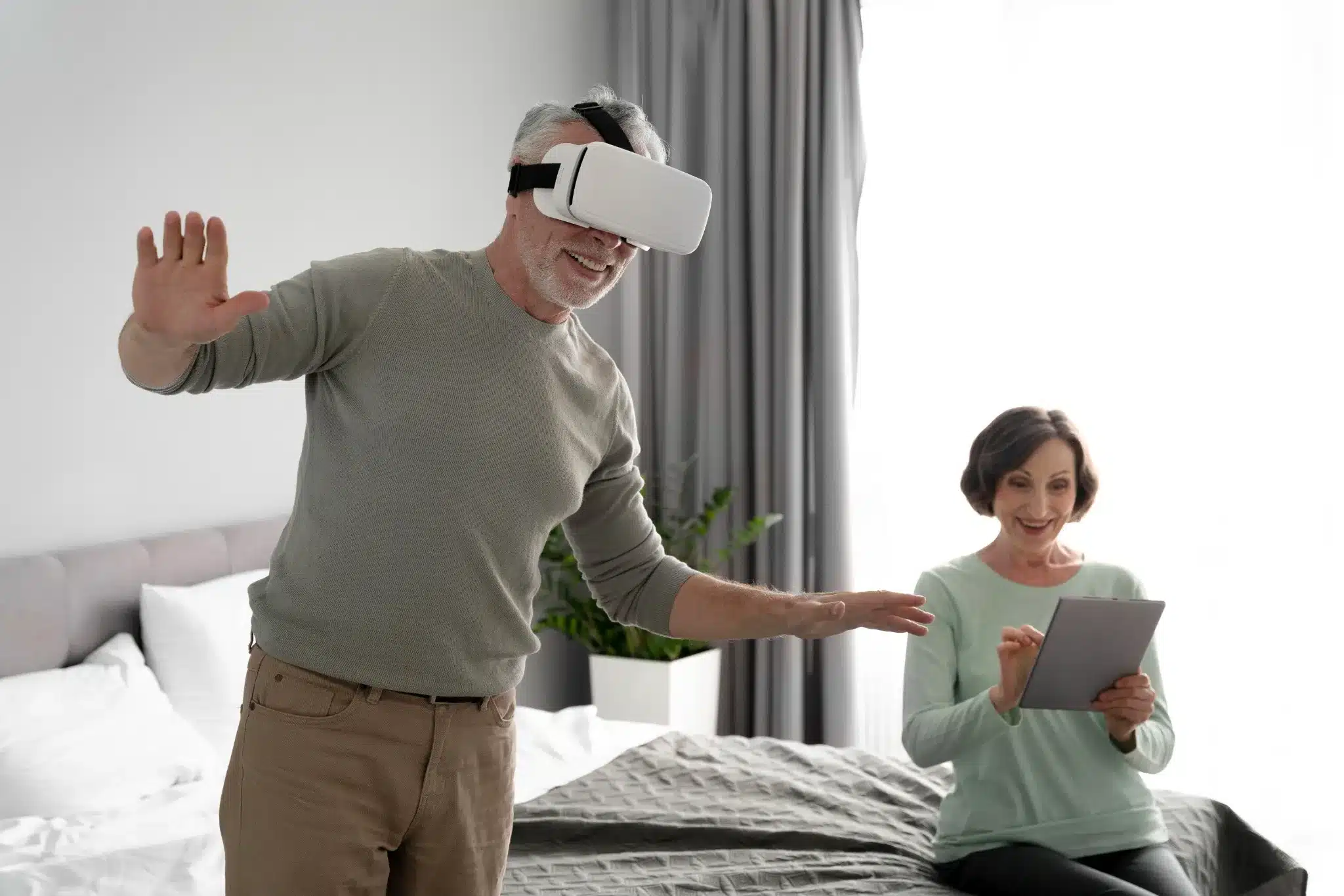 Jogos de realidade virtual para manter o corpo ativo Pessoa idosa jogando com óculos de realidade virtual enquanto outra assiste, usando um tablet.