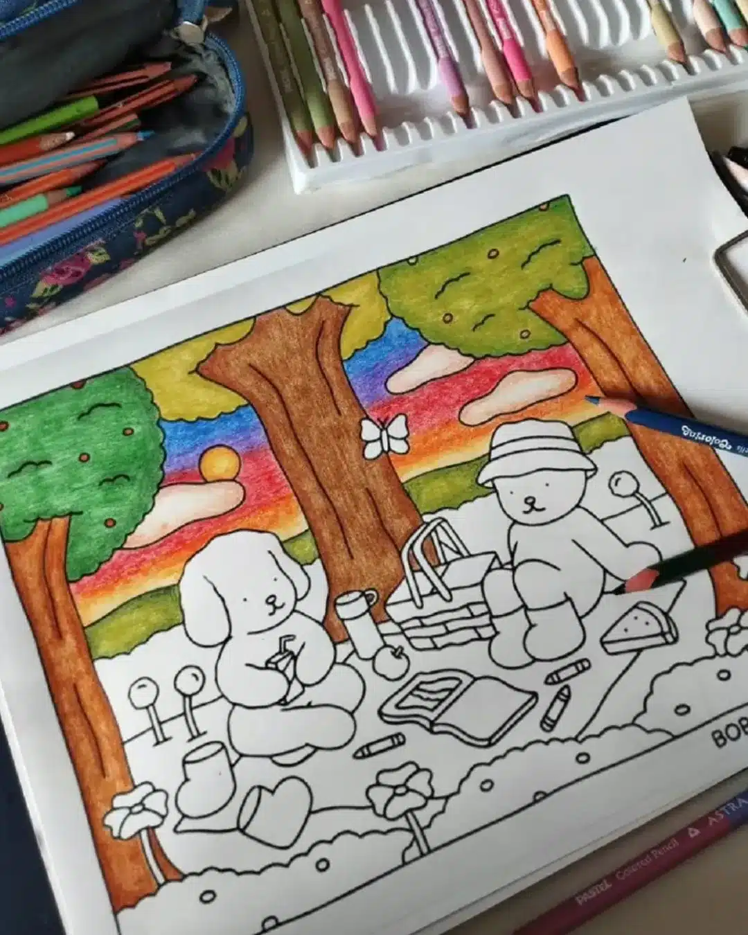 Ilustração de piquenique com personagens estilo Bobbie Goods, céu colorido ao fundo e lápis de cor ao redor.