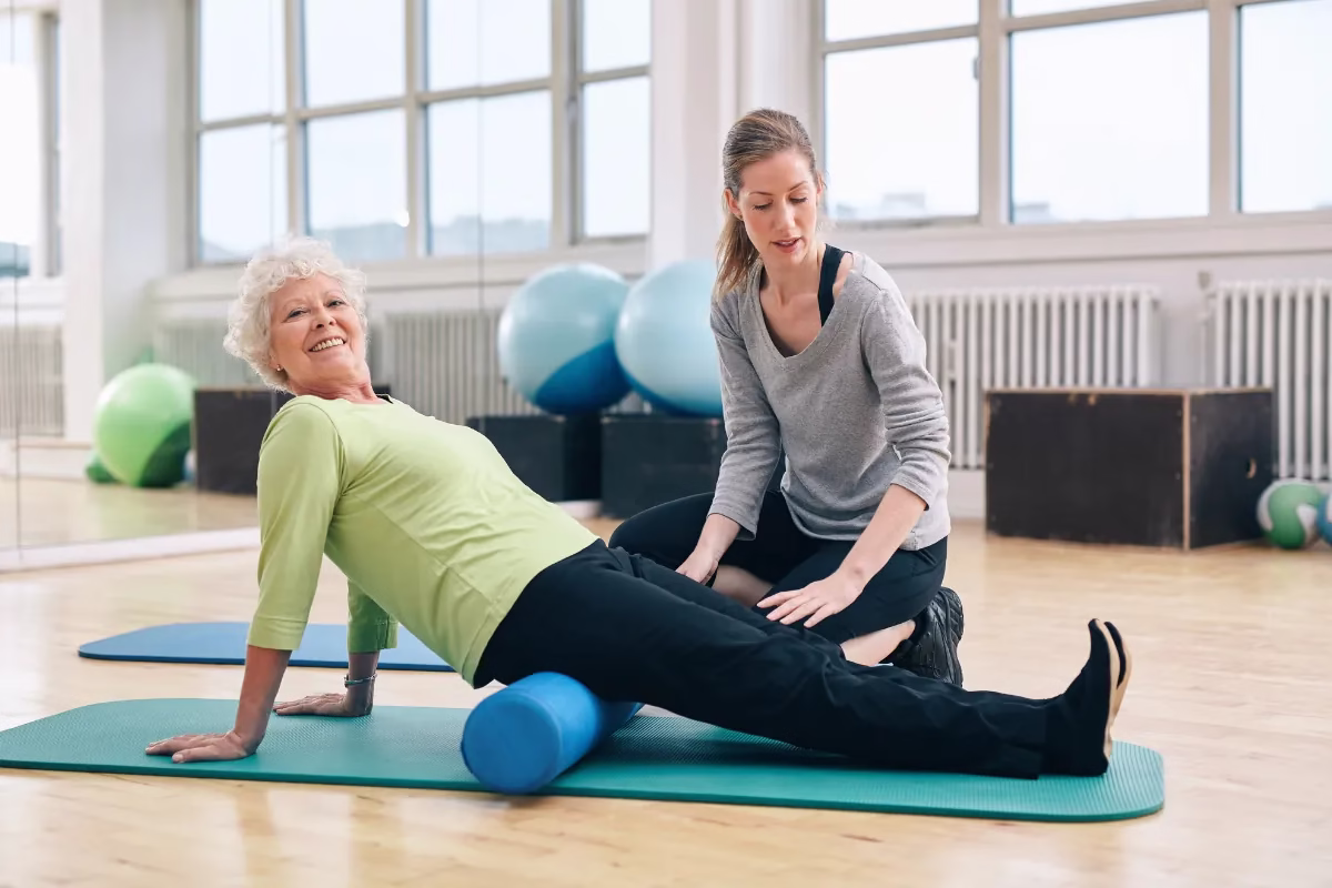 Idosa praticando pilates com auxílio de profissional em estúdio.