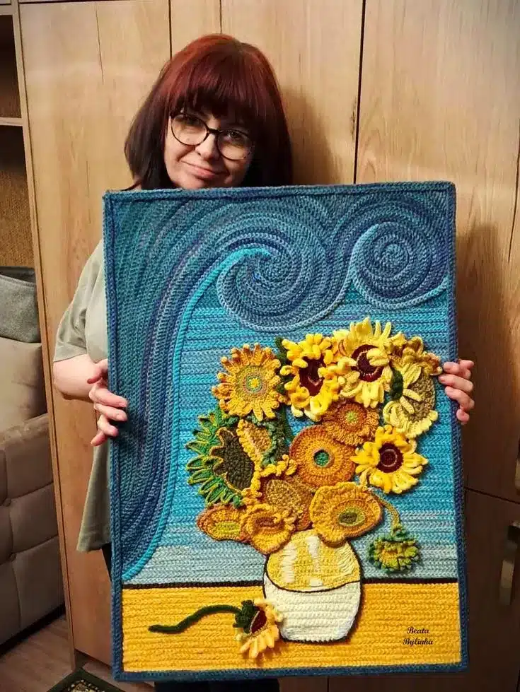 Quadro artístico de crochê com flores, representando um vaso de girassóis, em tons de amarelo e verde, sobre fundo colorido.