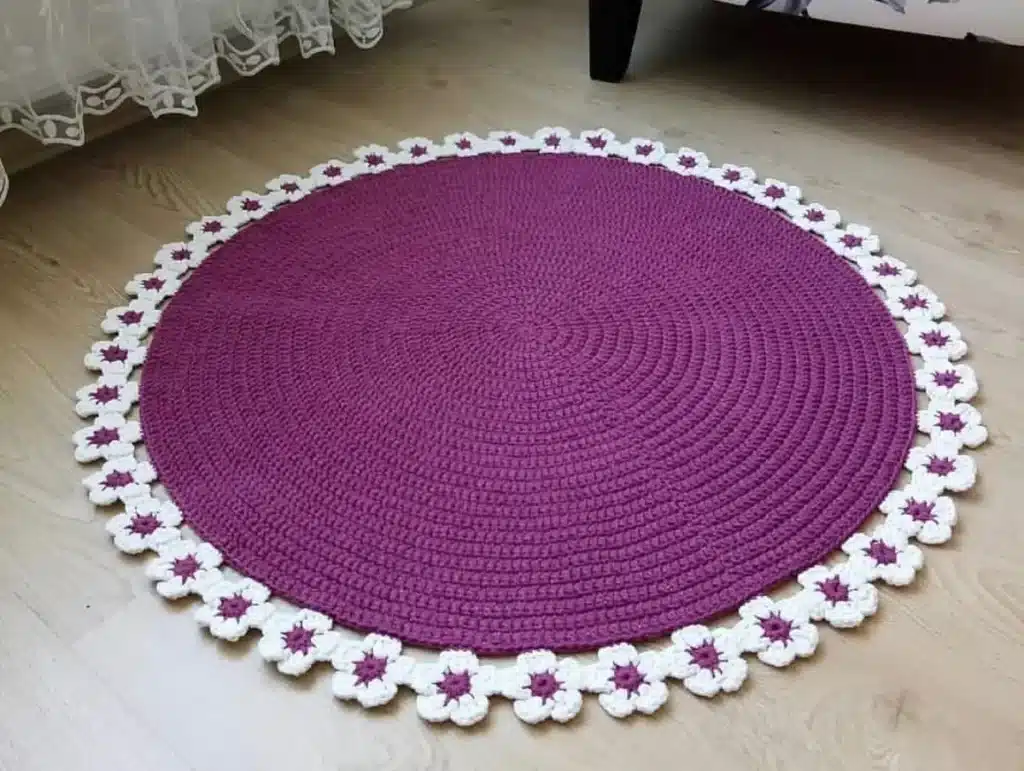 Tapete redondo de crochê com flores brancas e roxas ao redor.