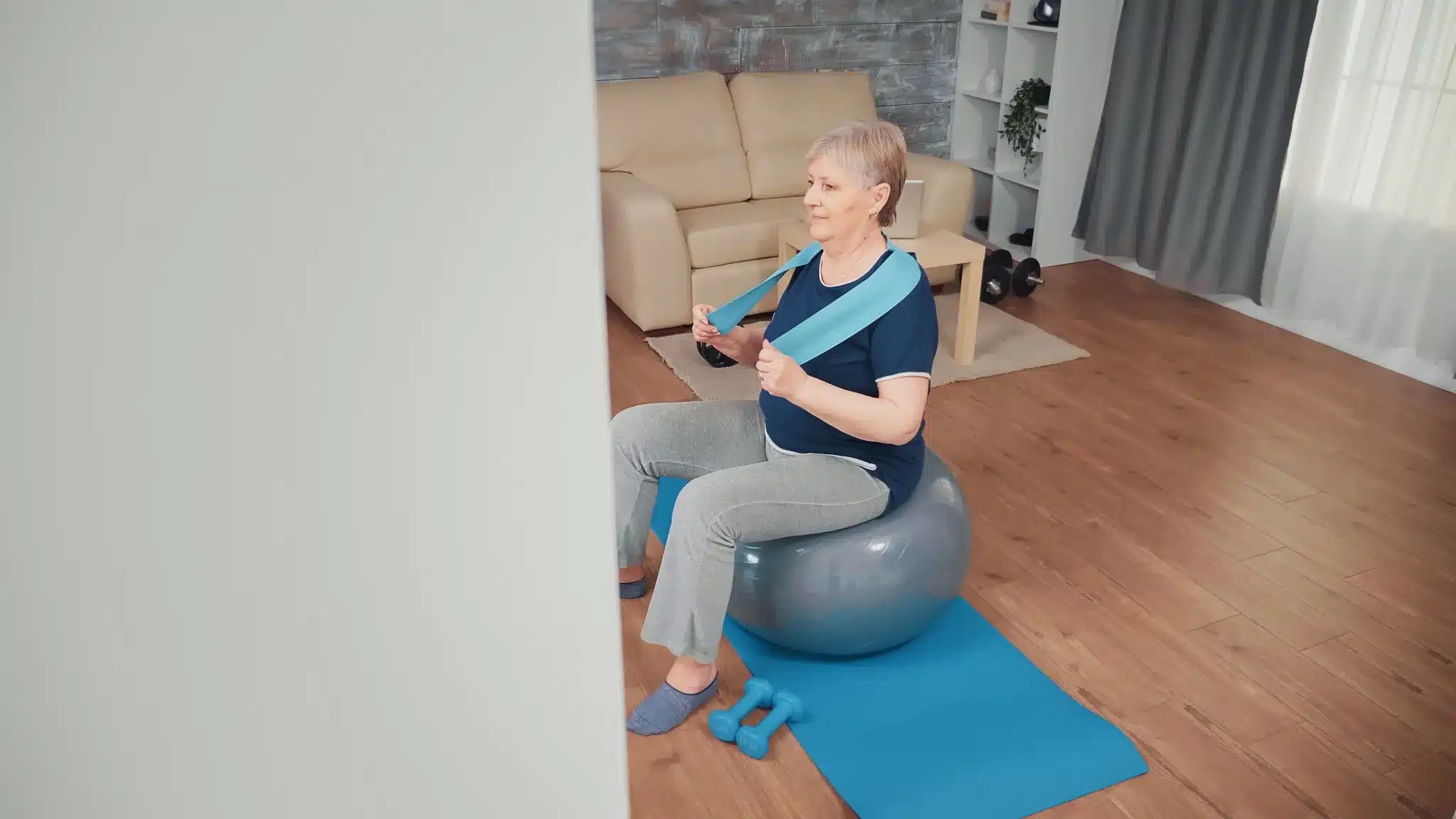 Idosa praticando exercícios com a bola suíça em casa, usando faixa elástica, para melhorar a saúde e mobilidade.