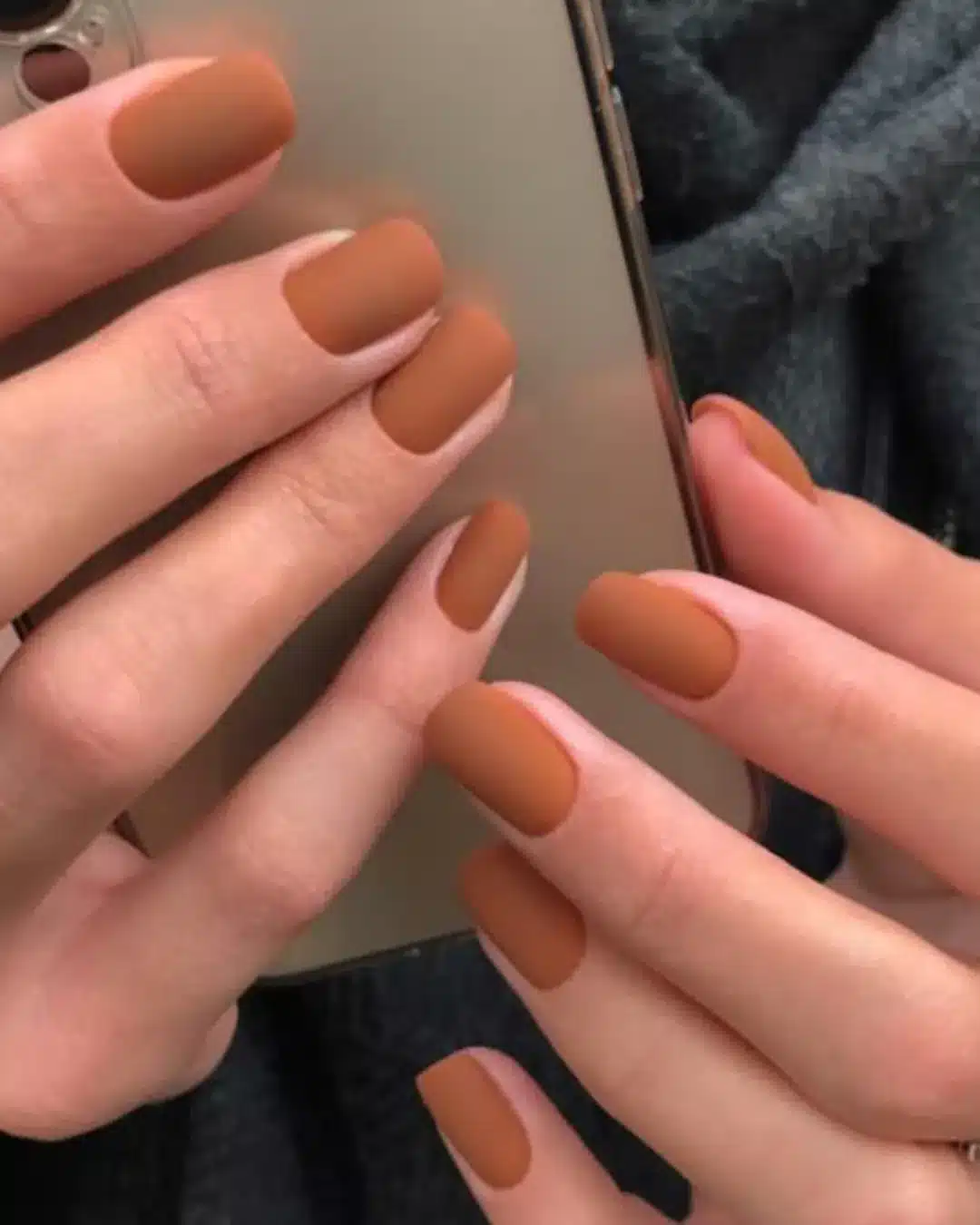 Unhas em tom terroso com acabamento fosco segurando celular