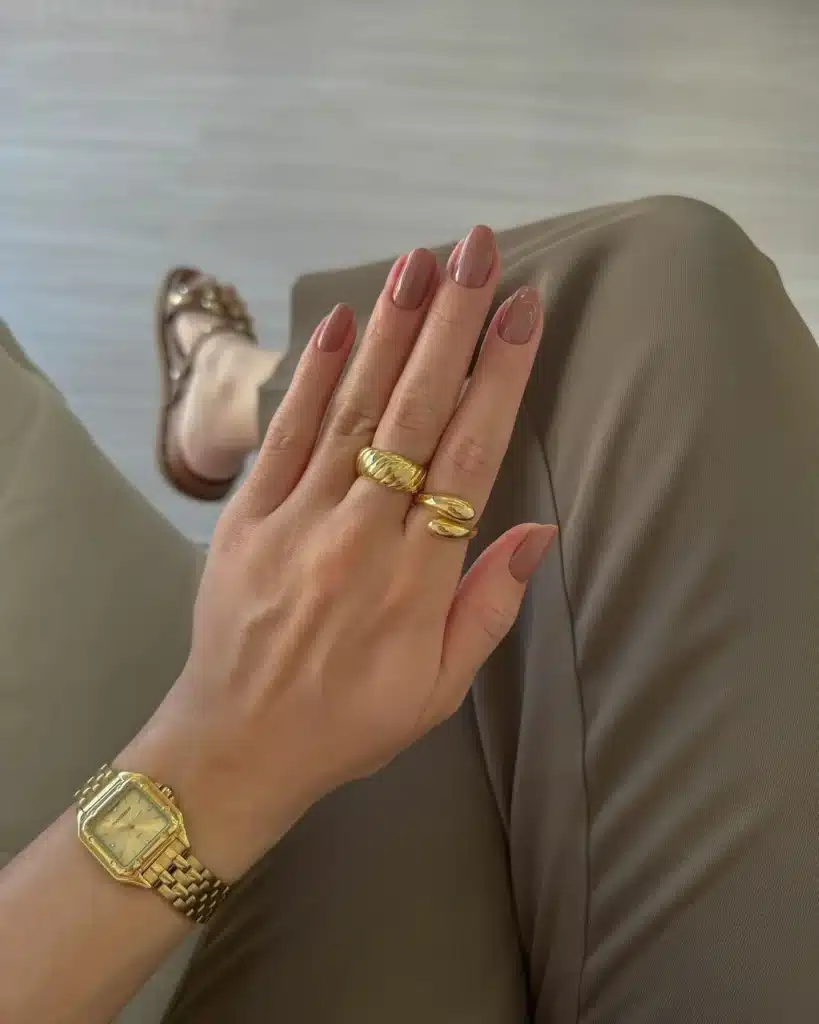 Imagem de unhas em tom nude, combinando com anéis dourados e relógio de pulso. O clássico tom nude é uma escolha elegante e versátil para qualquer ocasião.