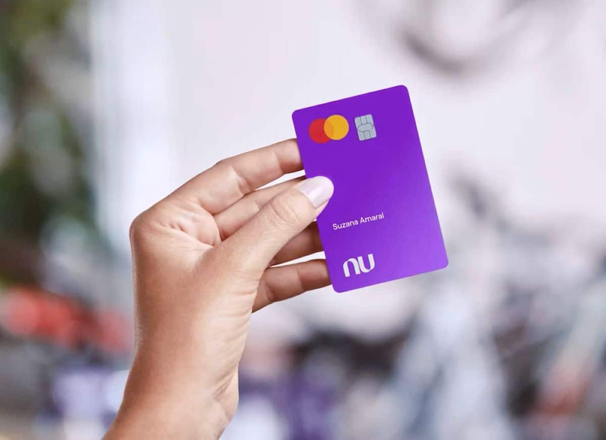 Mão segurando um cartão de crédito roxo do Nubank com o nome "Suzana Amaral" visível.
