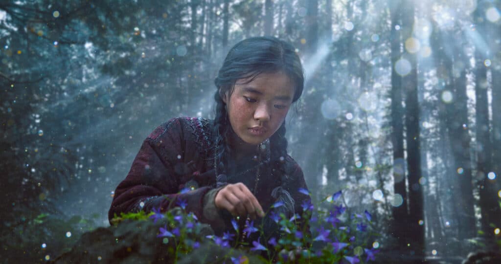 A protagonista do dorama "Kingdom" colhendo flores em uma floresta escura com raios de sol e pontos de luz.