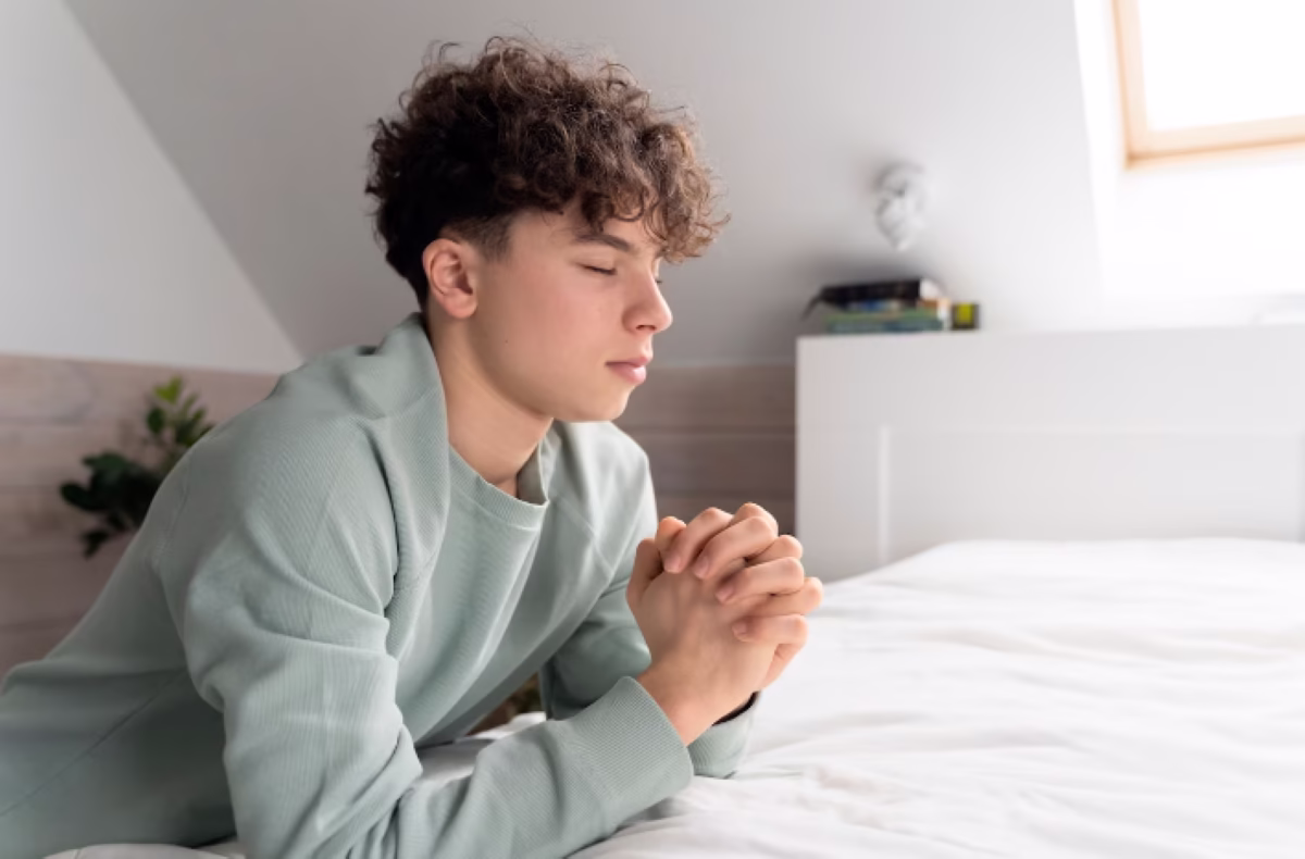 Jovem em oração ajoelhado ao lado da cama, simbolizando uma noite de paz e reflexão.