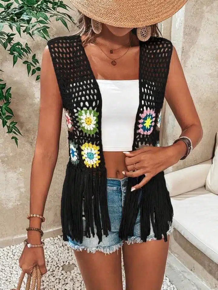 Imagem de um colete de crochê com quadrados granny coloridos em flores e franjas, com base preta, usado com shorts jeans.