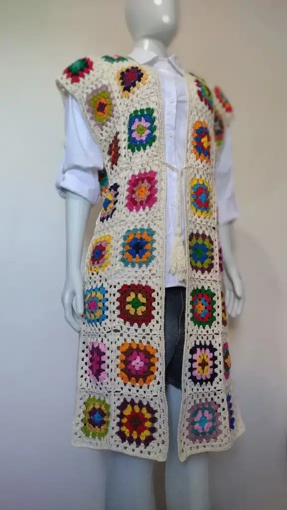 Imagem de um colete estilo granny square oversized, com quadrados coloridos e base clara, usado sobre uma camisa branca e shorts.