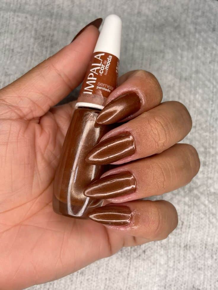 A imagem de uma mão segurando um frasco de esmalte de cor perolada, com as unhas pintadas em um tom de caramelo.