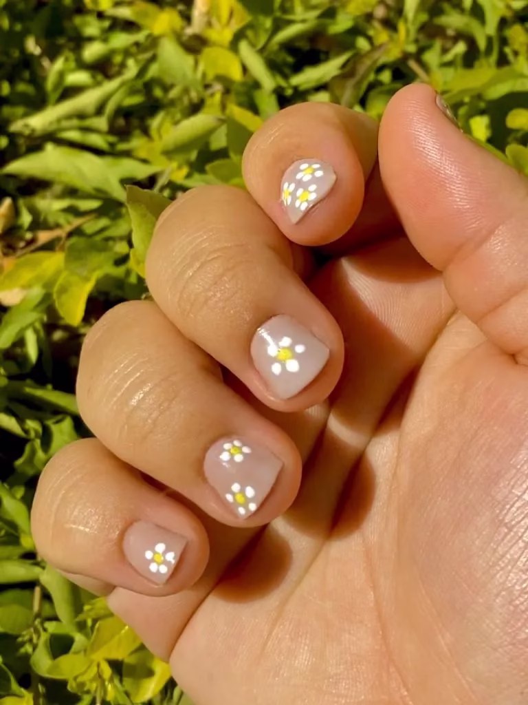 Unhas curtas com delicadas flores brancas e amarelas em fundo neutro.