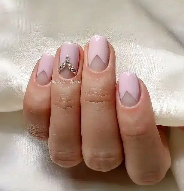 Unhas com francesinha invertida geométrica em tons pastéis de rosa, com detalhes em formato de 'V' e adornadas com cristais.
