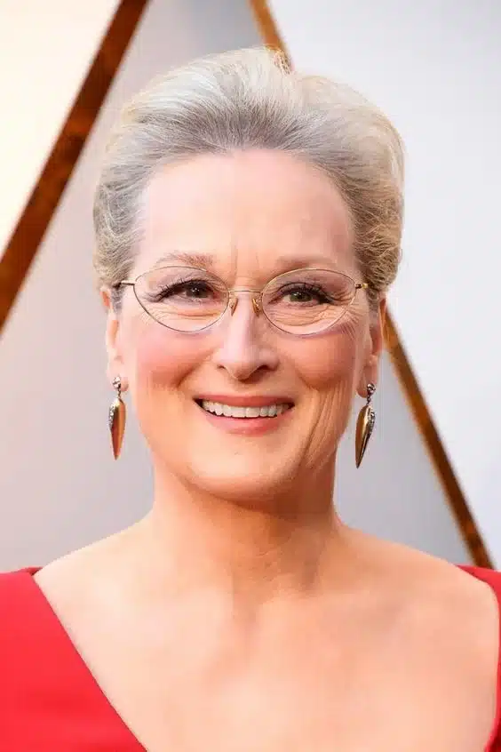 Meryl Streep sorrindo em um evento de tapete vermelho, usando óculos e brincos elegantes, com um vestido vermelho, destacando sua aparência confiante e sofisticada.