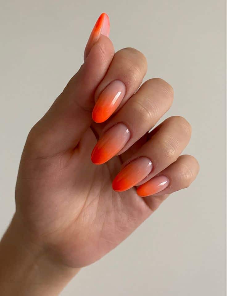 A imagem de uma mão com as unhas em formato amendoado, com esmalte em degradê que vai de um rosa claro na base a um laranja neon nas pontas.