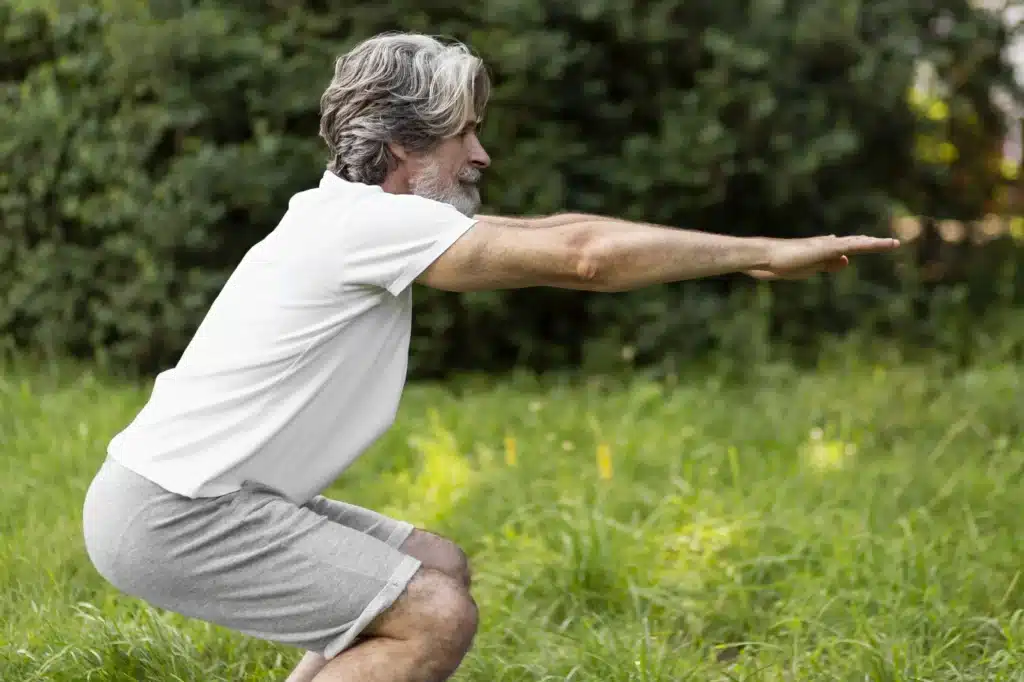 Homem idoso com barba e cabelo grisalhos, camiseta branca e shorts cinza, realizando agachamento leve ao ar livre em um gramado.