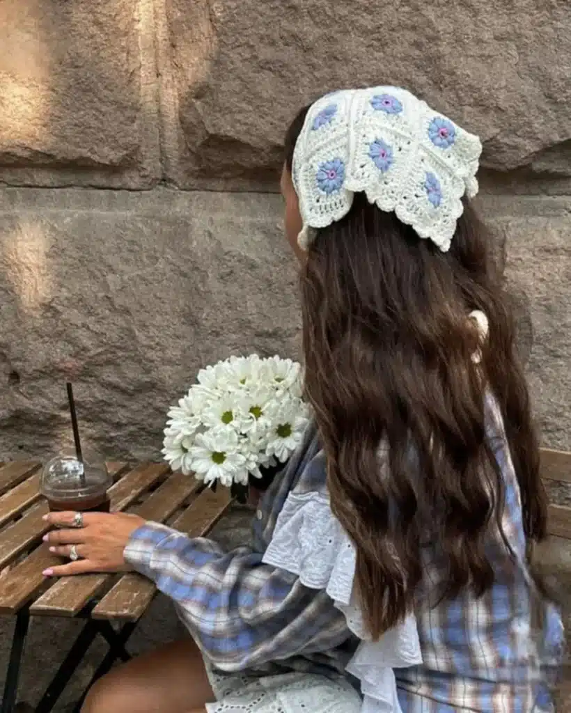 Mulher usando bandana de crochê com flores azuis e brancas, com cabelo longo e liso, segurando flores brancas enquanto desfruta de uma bebida.