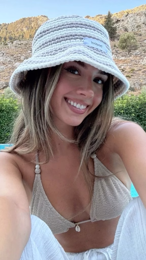 Mulher sorrindo com chapéu de crochê na praia, em frente à piscina.