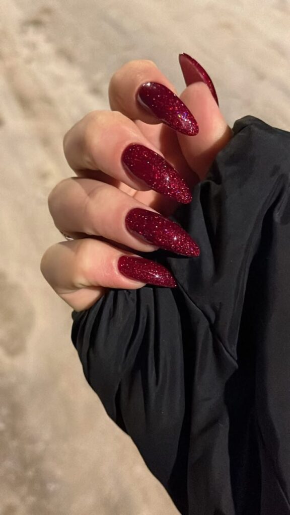 Mão com unhas stiletto longas pintadas de vermelho escuro com glitter.