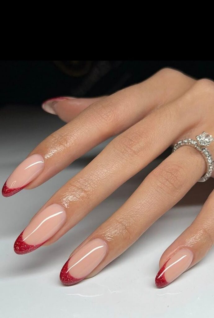 Unhas com um design de francesinha em formato stiletto, com as pontas decoradas em glitter vermelho.