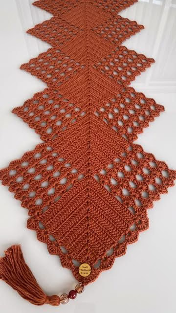  Trilho de mesa de crochê vermelho em formato geométrico com detalhes vazados e tassel decorativo na ponta.