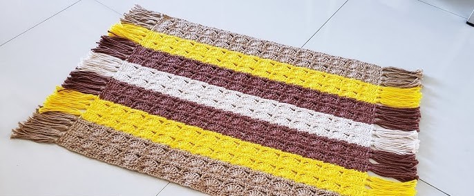 Um tapete de crochê retangular com listras coloridas em tons de marrom, bege e amarelo, com acabamento em tassel nas extremidades.