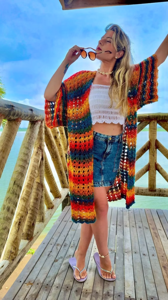 Mulher vestindo kimono de crochê colorido, óculos de sol e saia jeans, posando ao ar livre com vista para o mar.