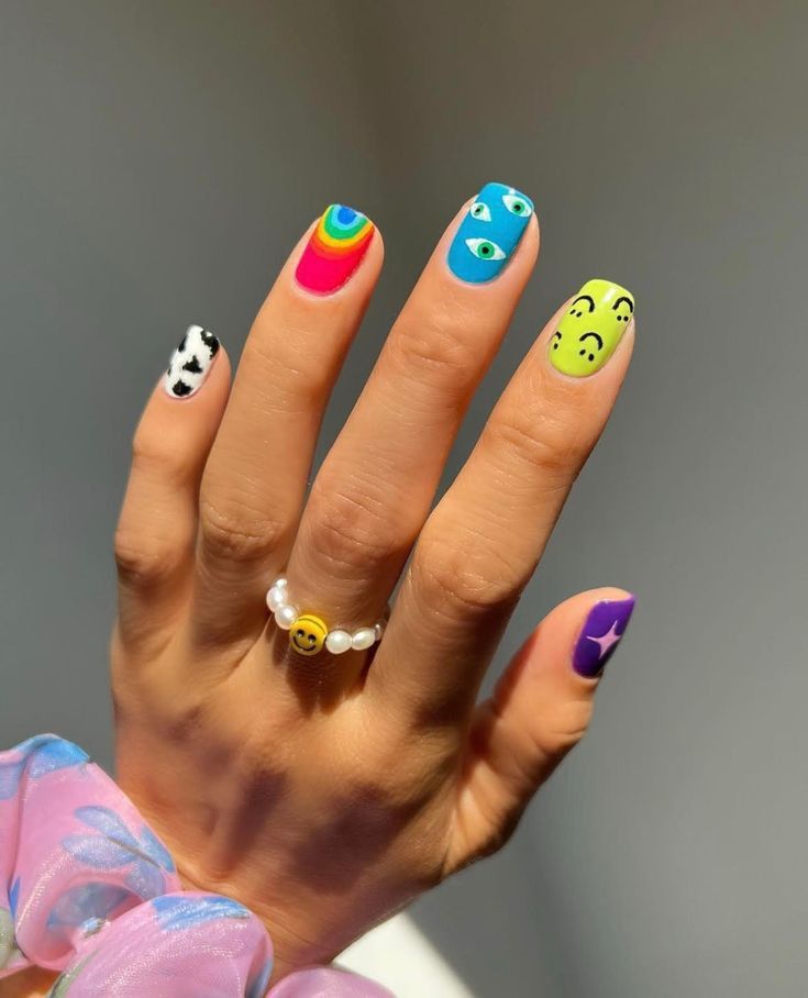 Mão mostrando unhas coloridas com nail art divertida: desenho de vaca, arco-íris, olhos, emojis e estrela.