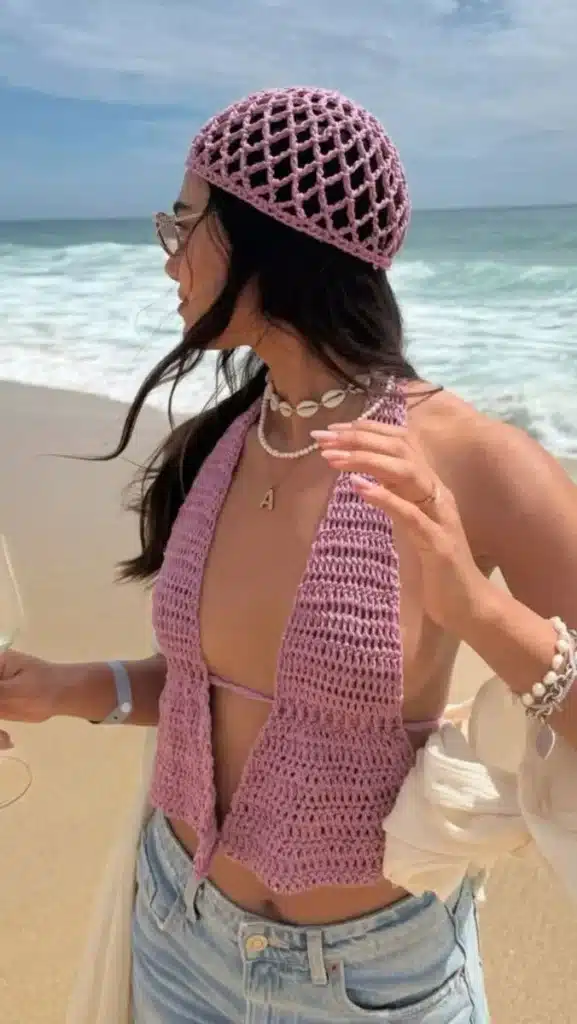 Mulher com look praiano usando touca de crochê rosa e colares, com o mar ao fundo.