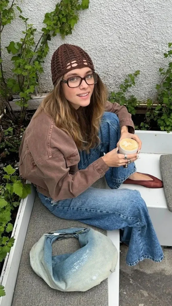 Mulher com look urbano despojado, usando touca de crochê marrom, jaqueta jeans e calça jeans.