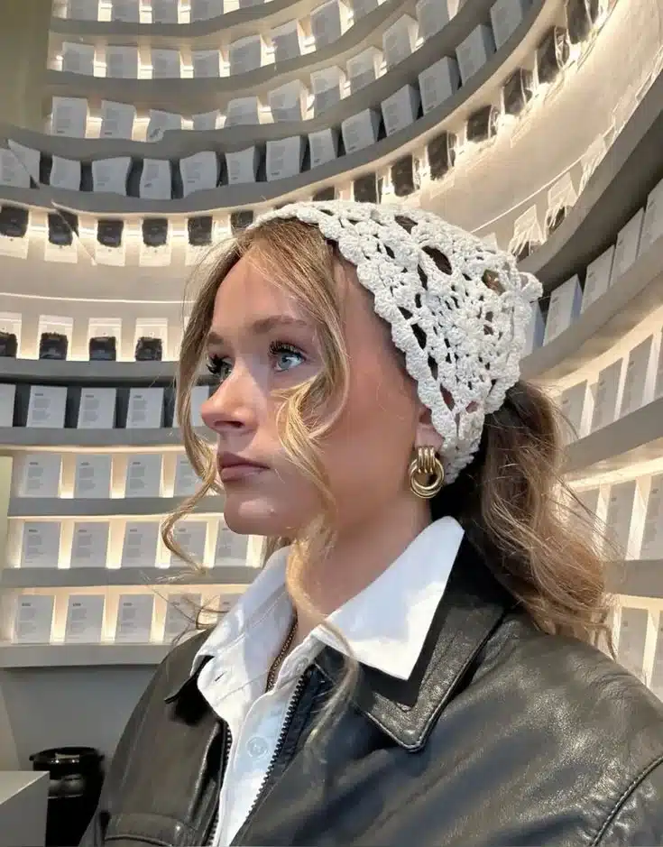 Mulher usando touca de crochê branca, blusa de botão branca e jaqueta de couro preta, com um ambiente sofisticado ao fundo.