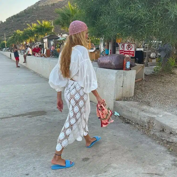 Mulher caminhando com look urbano despojado, usando touca de crochê rosa e vestido branco de crochê.