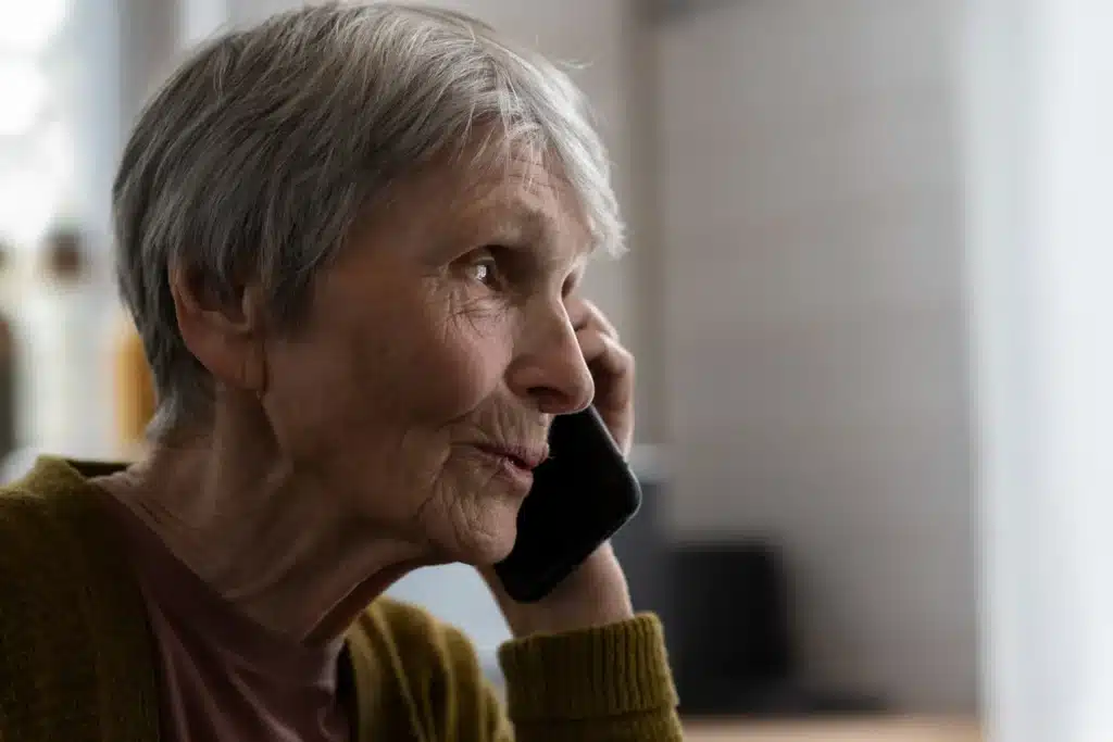 Mulher idosa falando ao telefone, com expressão atenta, em ambiente doméstico.
